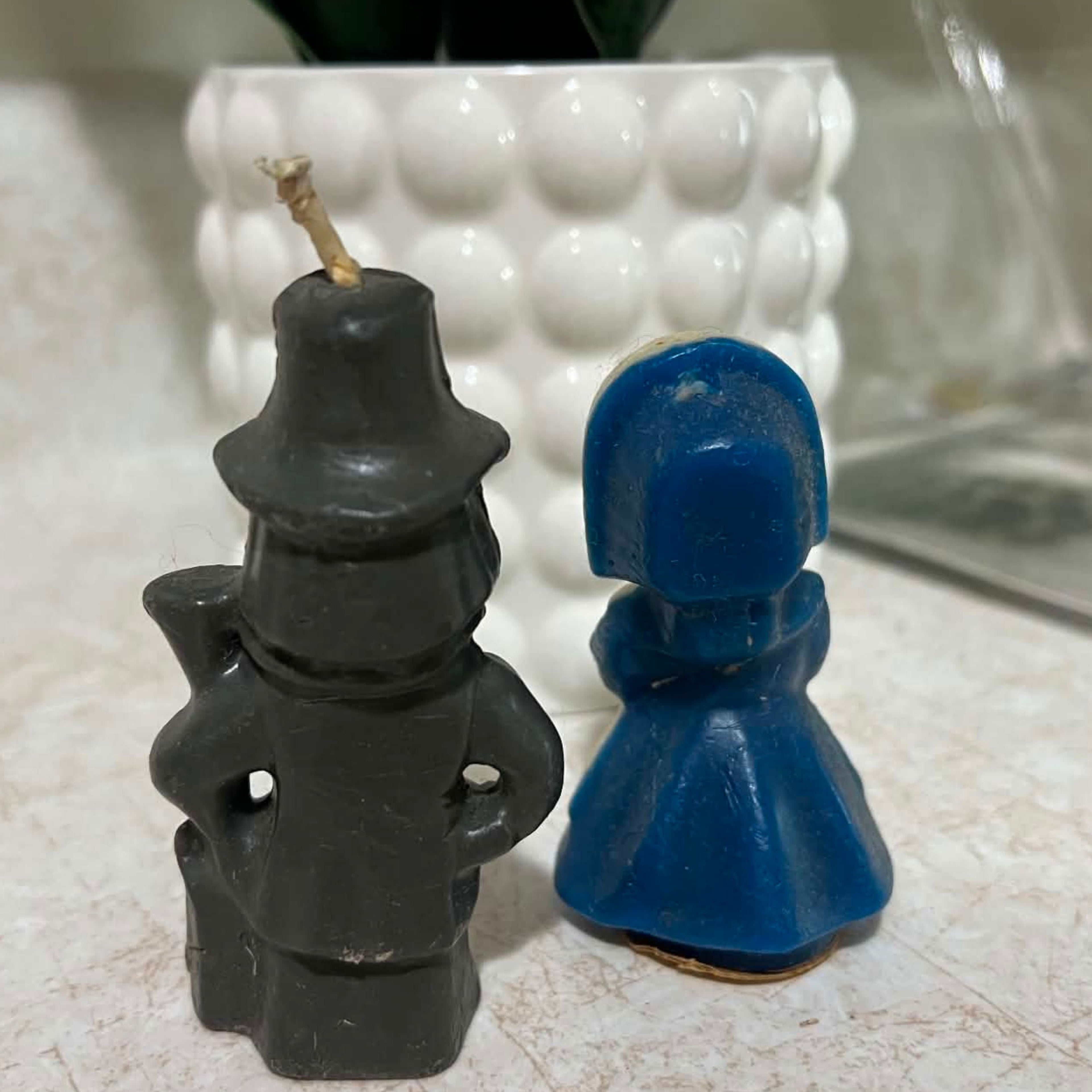 Back view of two vintage Gurley Thanksgiving candles showing the charcoal wax clothing and long gun on the male pilgrim and deep blue molded bonnet and dress on the female figure. Both candles display surface wear and marks consistent with age and storage.