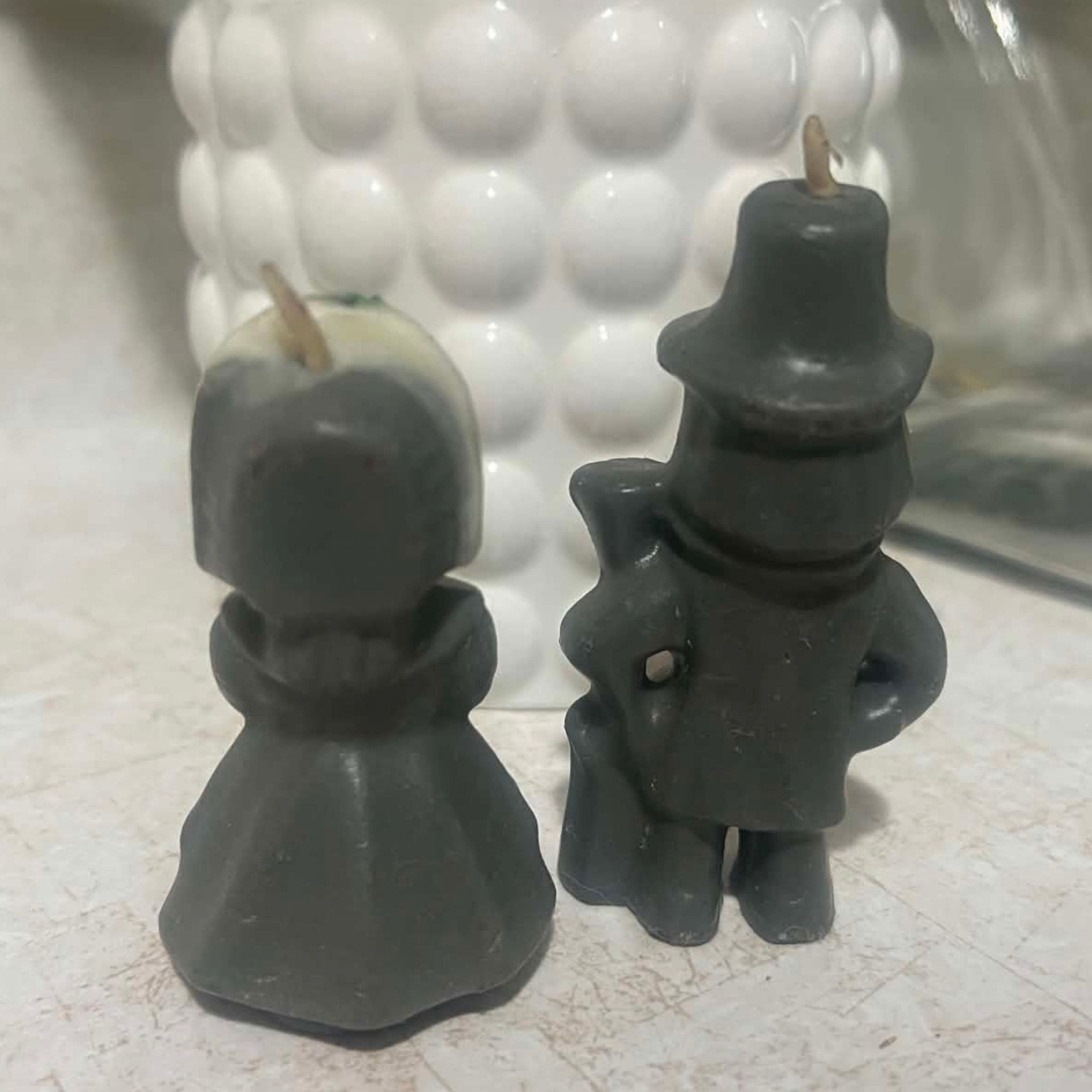 Back view of a vintage Gurley Thanksgiving pilgrim candle set showing the dark charcoal wax clothing, molded bonnet, pilgrim hat and back of the male figure's long gun. Both small figural candles retain their visible wicks and show age-related surface wear.