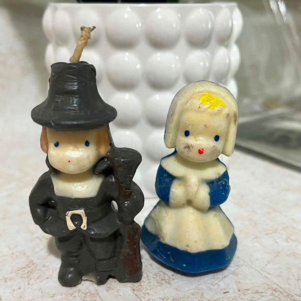 Vintage Gurley Thanksgiving pilgrim candle pair shown from the front, with a male figure in dark clothing and hat beside a female pilgrim in a blue and cream dress. Both molded wax figures have painted blue eyes and red mouths with visible age-related wear.