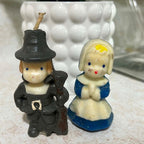 Vintage Gurley Thanksgiving pilgrim candle pair shown from the front, with a male figure in dark clothing and hat beside a female pilgrim in a blue and cream dress. Both molded wax figures have painted blue eyes and red mouths with visible age-related wear.