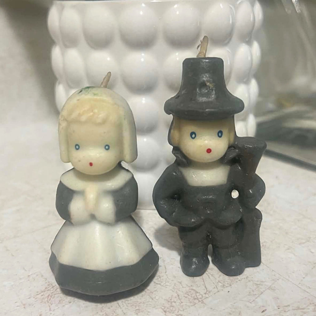 Vintage Gurley Thanksgiving pilgrim candle pair shown from the front, featuring a bonneted woman with clasped hands and a pilgrim man in a dark hat carrying a long gun. Both molded wax candles have cream faces, blue eyes, red mouths and intact unused wicks.