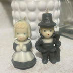 Vintage Gurley Thanksgiving pilgrim candle pair shown from the front, featuring a bonneted woman with clasped hands and a pilgrim man in a dark hat carrying a long gun. Both molded wax candles have cream faces, blue eyes, red mouths and intact unused wicks.