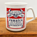 Front view of a vintage 1990s Jamaica souvenir coffee mug featuring a bold red and white beer label-inspired graphic with playful typography celebrating Jamaica as the King of Beaches. The ceramic travel mug sits on a rustic wooden surface with its colorful vacation-themed artwork fully visible.