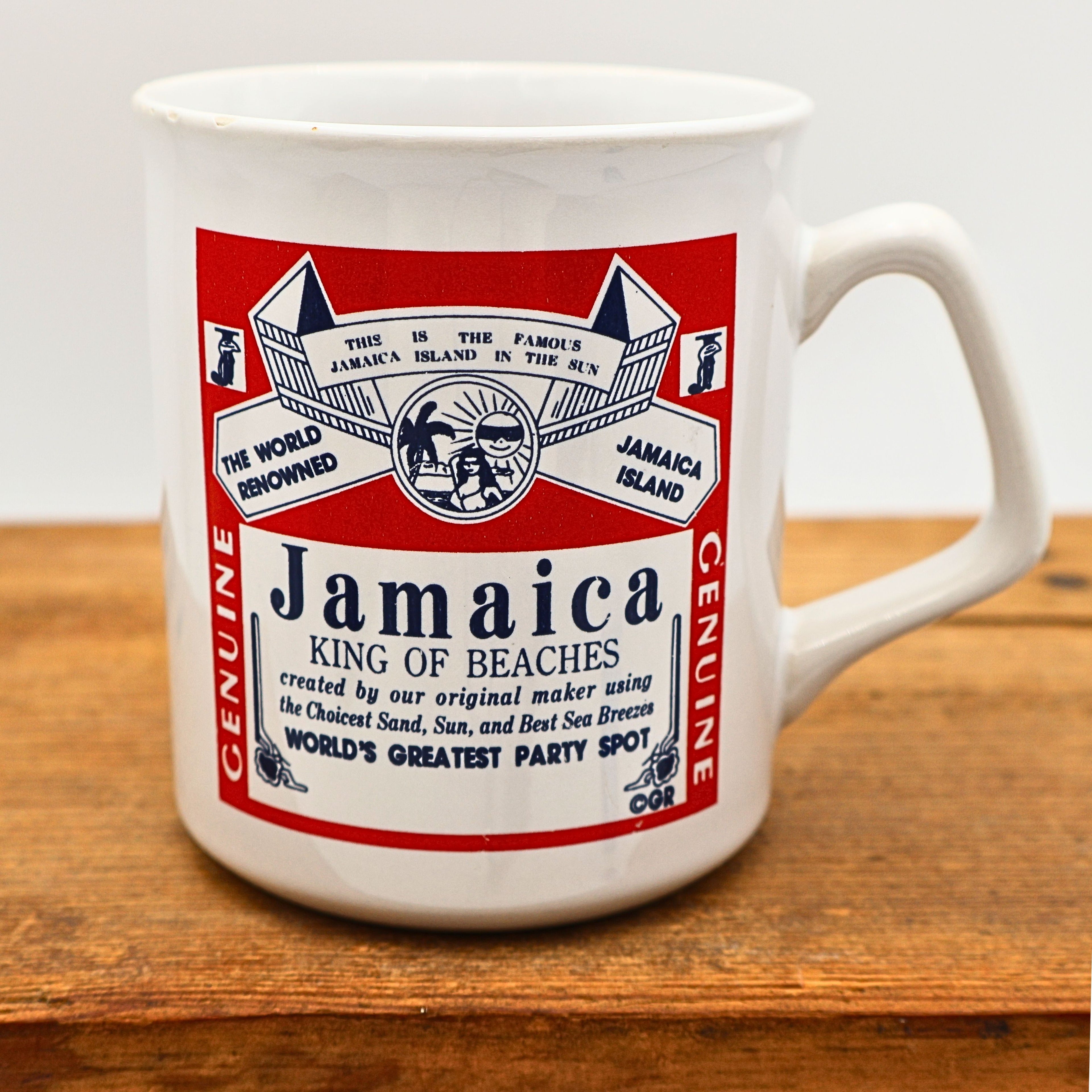 Front view of a vintage 1990s Jamaica souvenir coffee mug featuring a bold red and white beer label-inspired graphic with playful typography celebrating Jamaica as the King of Beaches. The ceramic travel mug sits on a rustic wooden surface with its colorful vacation-themed artwork fully visible.