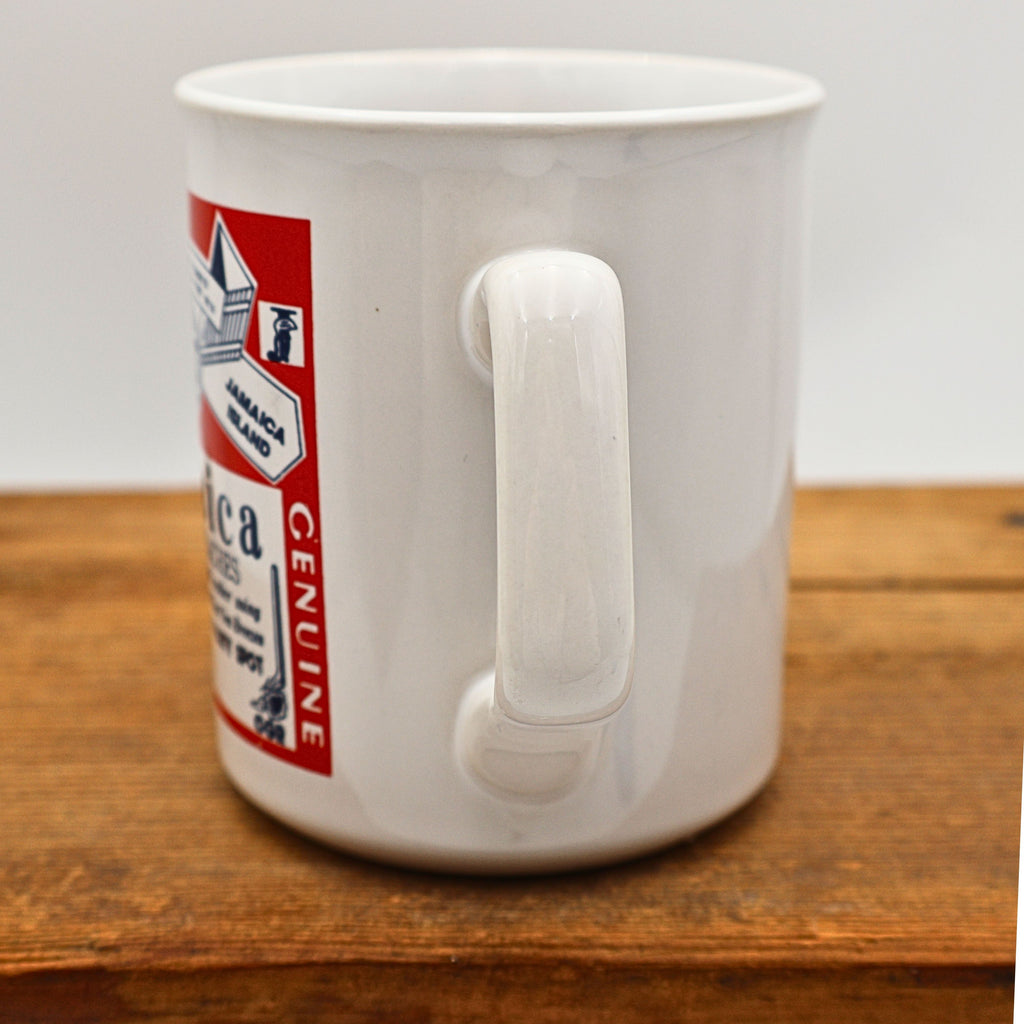 Side view showing the sturdy handle of the vintage Jamaica souvenir ceramic mug. Part of the colorful red travel graphic wraps around the front while the glossy white glaze and classic cylindrical shape remain clearly visible against a neutral background.