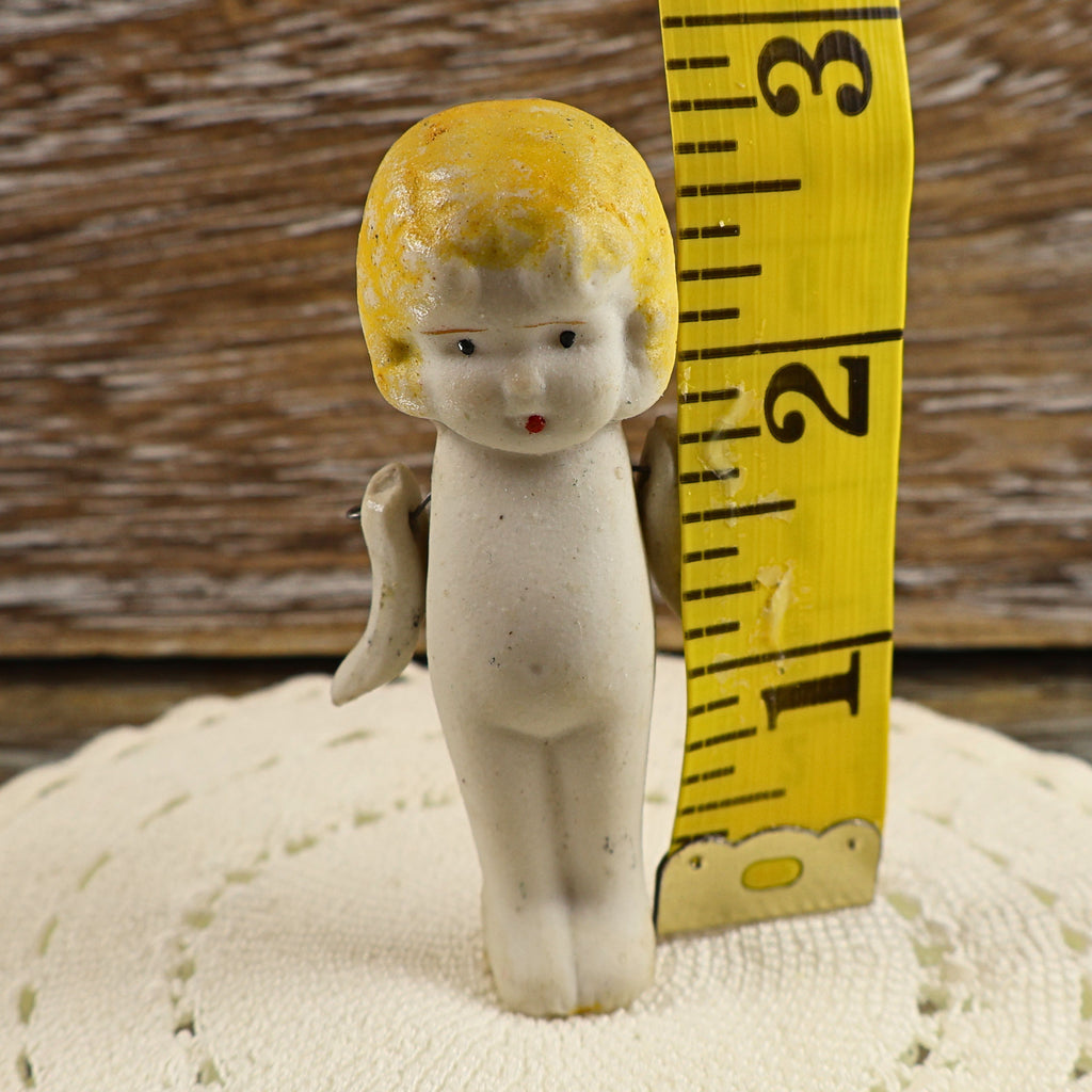 Front view of the vintage Japanese bisque penny doll beside a yellow measuring tape indicating a height of approximately two and three-quarter inches. Her molded blonde hair, painted face, petite porcelain body, movable arms, and narrow legs are clearly visible.