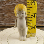 Front view of the vintage Japanese bisque penny doll beside a yellow measuring tape indicating a height of approximately two and three-quarter inches. Her molded blonde hair, painted face, petite porcelain body, movable arms, and narrow legs are clearly visible.