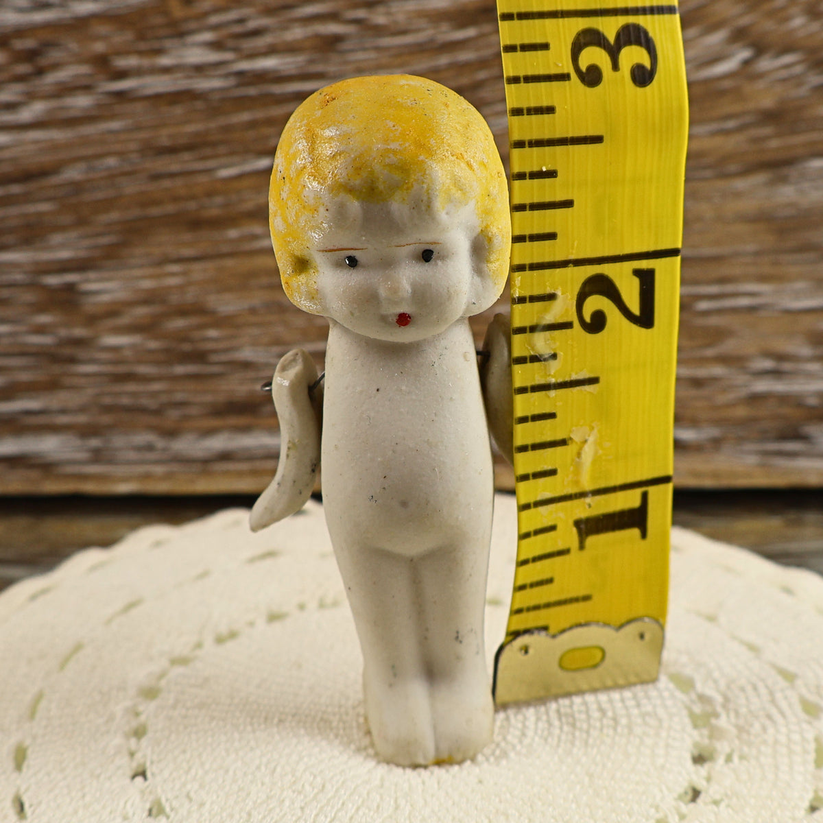 Front view of the vintage Japanese bisque penny doll beside a yellow measuring tape indicating a height of approximately two and three-quarter inches. Her molded blonde hair, painted face, petite porcelain body, movable arms, and narrow legs are clearly visible.