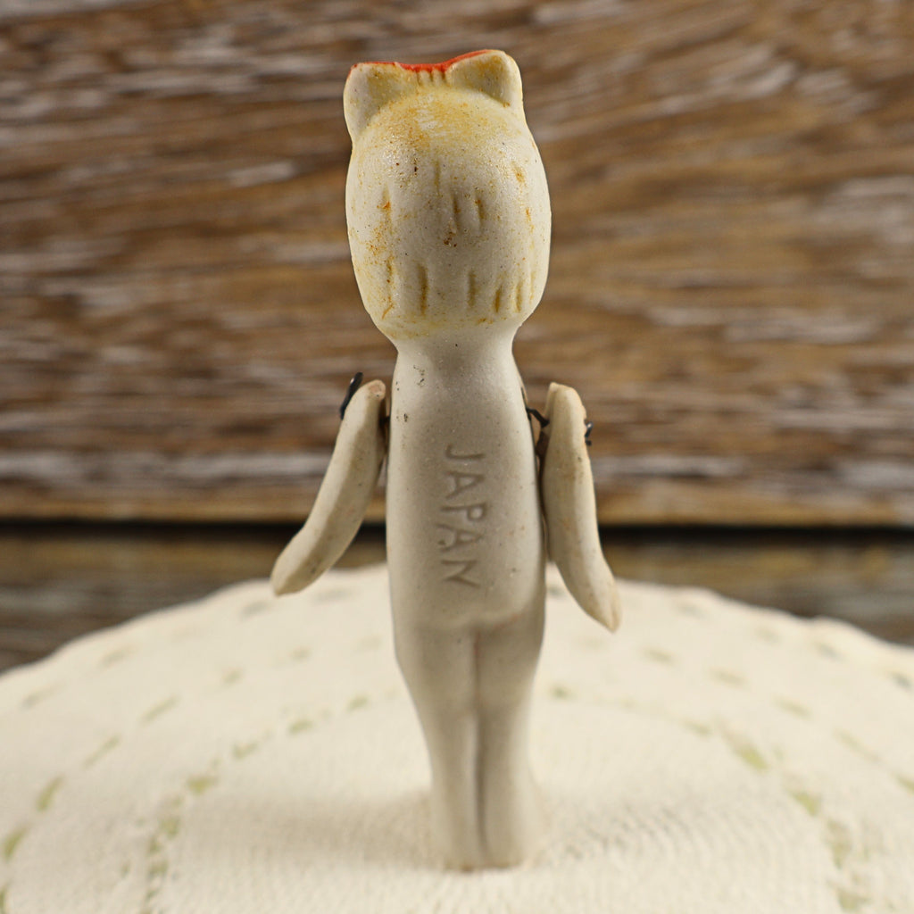 Back view of a small vintage bisque doll showing the vertically impressed JAPAN country-of-origin mark along her torso. Her molded hair, faded red bow, stationary legs, and two wire-jointed arms are visible against cream needlework and a weathered wooden backdrop.