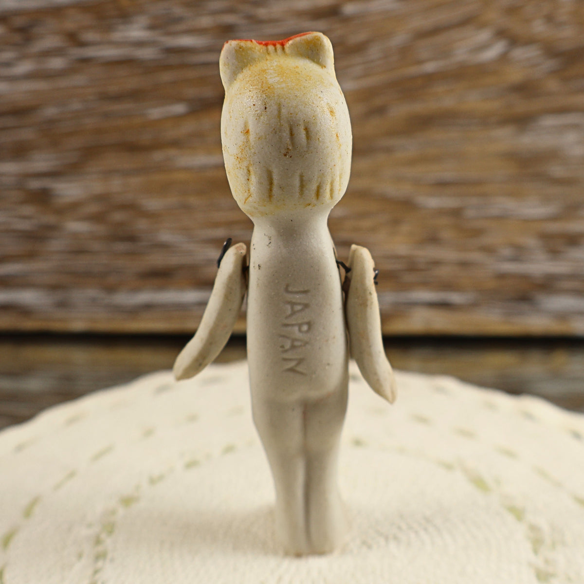 Back view of a small vintage bisque doll showing the vertically impressed JAPAN country-of-origin mark along her torso. Her molded hair, faded red bow, stationary legs, and two wire-jointed arms are visible against cream needlework and a weathered wooden backdrop.