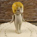 Front view of a petite vintage Japanese bisque penny doll standing on an ivory crocheted textile. The miniature figure has molded golden-yellow bobbed hair, painted dark eyes, red lips, an unglazed ivory body, and two separately attached movable arms.