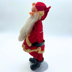 Left-facing profile of the vintage Japanese Santa doll, showing his projecting molded nose, long white beard, red fabric cap, and yellowed trim. A black belt circles the suit, while the metal support and gray circular base hold the figure above his black boots.