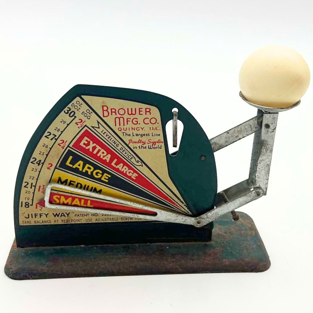 Vintage Brower-branded Jiffy-Way egg scale photographed with a pale egg resting in the raised metal cup. The front chart shows colored size bands and ounce markings, while the open pointer crosses its lower portion above a worn and rusted green base.