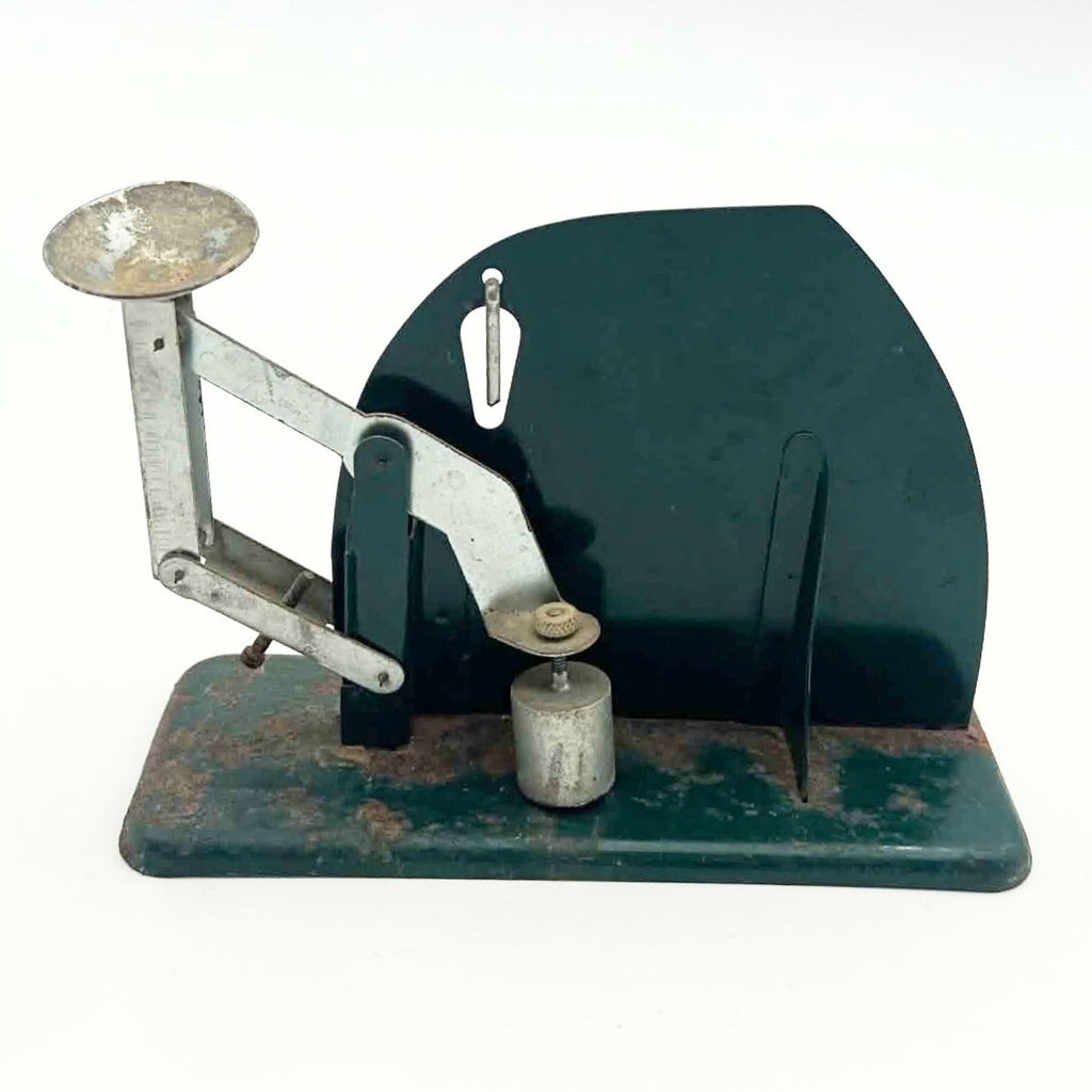 Rear view of a vintage Jiffy-Way egg scale with a dark green panel, metal balance arms and an elevated egg cup at the left. A cylindrical counterweight sits beneath an adjustment screw near the center, above a base showing widespread rust and paint loss.