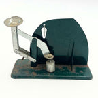 Rear view of a vintage Jiffy-Way egg scale with a dark green panel, metal balance arms and an elevated egg cup at the left. A cylindrical counterweight sits beneath an adjustment screw near the center, above a base showing widespread rust and paint loss.