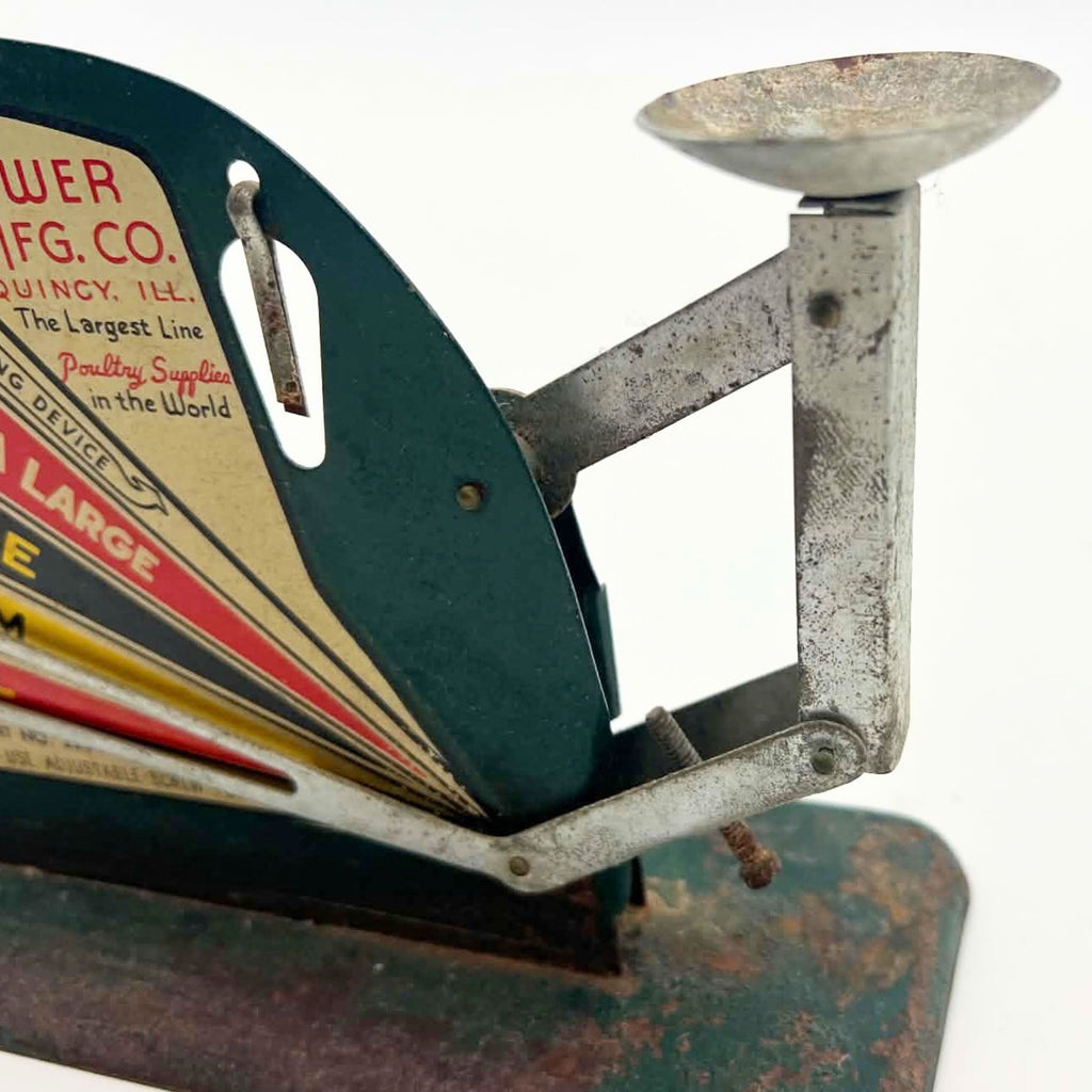 Close-up of a vintage Jiffy-Way egg scale showing the shallow metal egg cup, upright support and linked balance arms. Part of the Brower graphic panel and suspended leveling pin appear beside mottled metal surfaces and a green base with rust and paint loss.