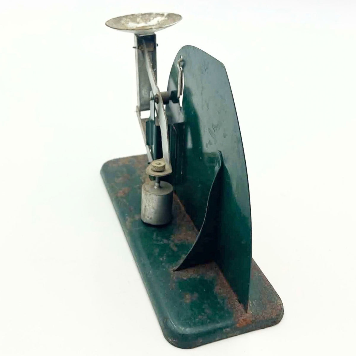 Narrow rear-angle view of a Jiffy-Way egg scale showing the green back panel, support bracket and linked metal mechanism. An adjustable cylindrical counterweight hangs near the worn base, while the elevated egg cup and suspended leveling pin remain visible.