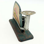 Side-angle view of a vintage Jiffy-Way egg scale on a white background, with its shallow round cup and vertical metal support prominent at the right. The chart is seen edge-on beside the balance linkage, above a green base with brown rust and paint loss.