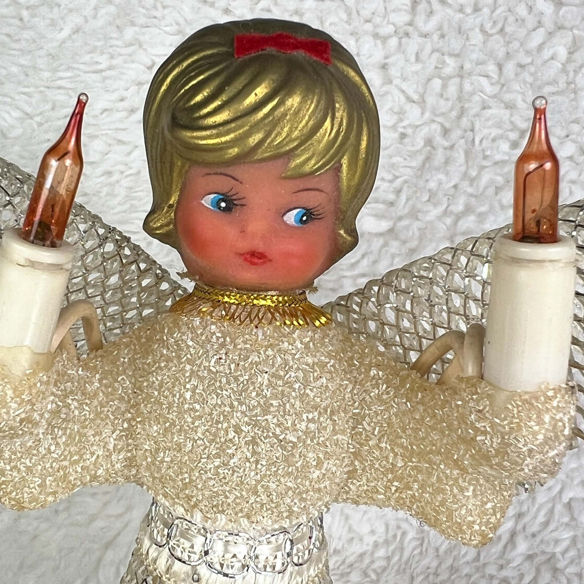 Close-up of a vintage angel tree topper with painted blue eyes, gold-colored molded hair, and a red bow. Two unlit amber flame-shaped bulbs rise beside her face, with cream candle holders, a textured bodice, gold neckline trim, and silver mesh wings visible.