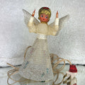 Unlit vintage angel tree topper standing on a reflective surface, with a cream textured bodice, silver mesh skirt and wings, gold-colored hair, and a red bow. Two amber candle bulbs are raised beside her face; the ivory cord, plug, and red lighting tag sit nearby.