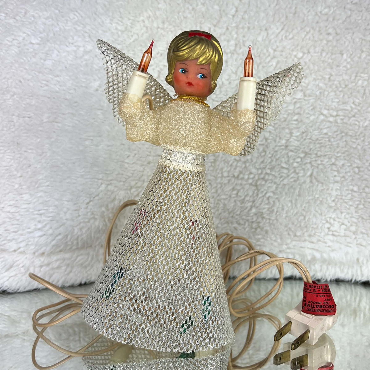 Unlit vintage angel tree topper standing on a reflective surface, with a cream textured bodice, silver mesh skirt and wings, gold-colored hair, and a red bow. Two amber candle bulbs are raised beside her face; the ivory cord, plug, and red lighting tag sit nearby.