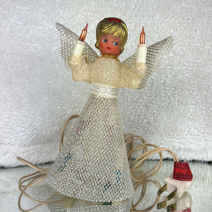 Unlit vintage angel tree topper standing on a reflective surface, with a cream textured bodice, silver mesh skirt and wings, gold-colored hair, and a red bow. Two amber candle bulbs are raised beside her face; the ivory cord, plug, and red lighting tag sit nearby.