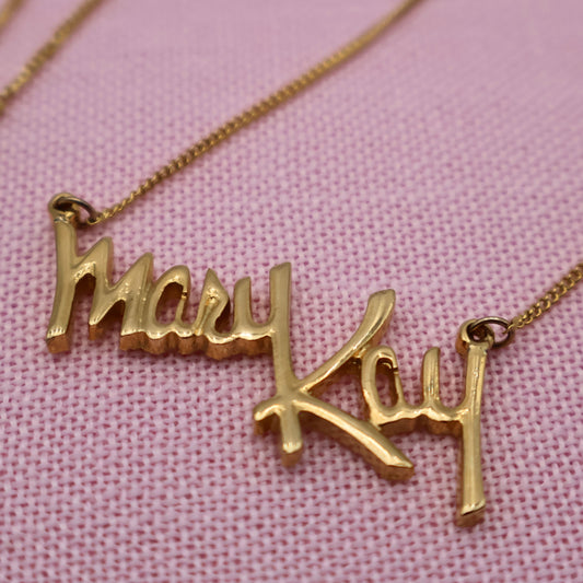 Vintage Mary Kay necklace in gold tone script by Sarah Coventry, close-up of signature pendant on pink background.