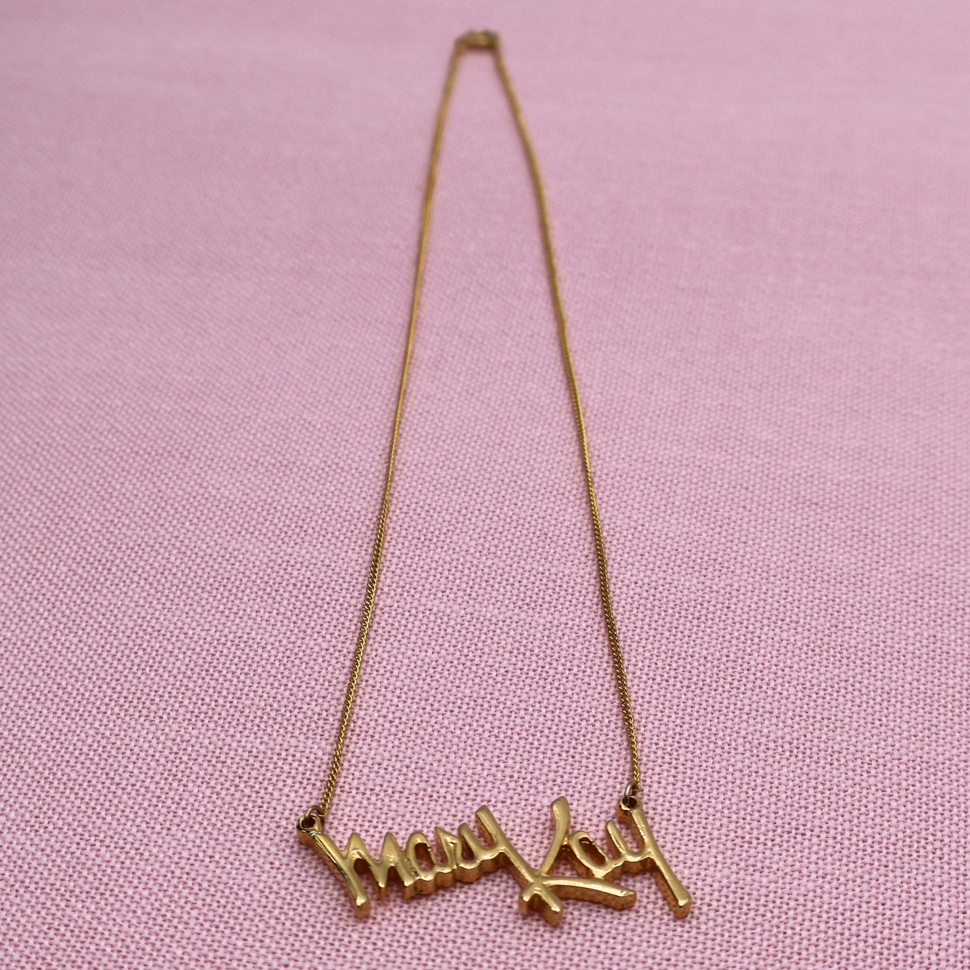 Full length view of vintage Mary Kay signature necklace with gold tone chain and script pendant on pink fabric background.