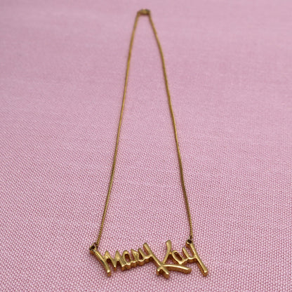Full length view of vintage Mary Kay signature necklace with gold tone chain and script pendant on pink fabric background.