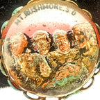 Close-up photograph of a vintage Mount Rushmore clip-on earring highlighting the detailed raised presidential portraits beneath the clear dome. The colorful scenic finish, gold-tone setting, and dimensional relief illustrate the artistry commonly found in mid-century South Dakota souvenir jewelry.