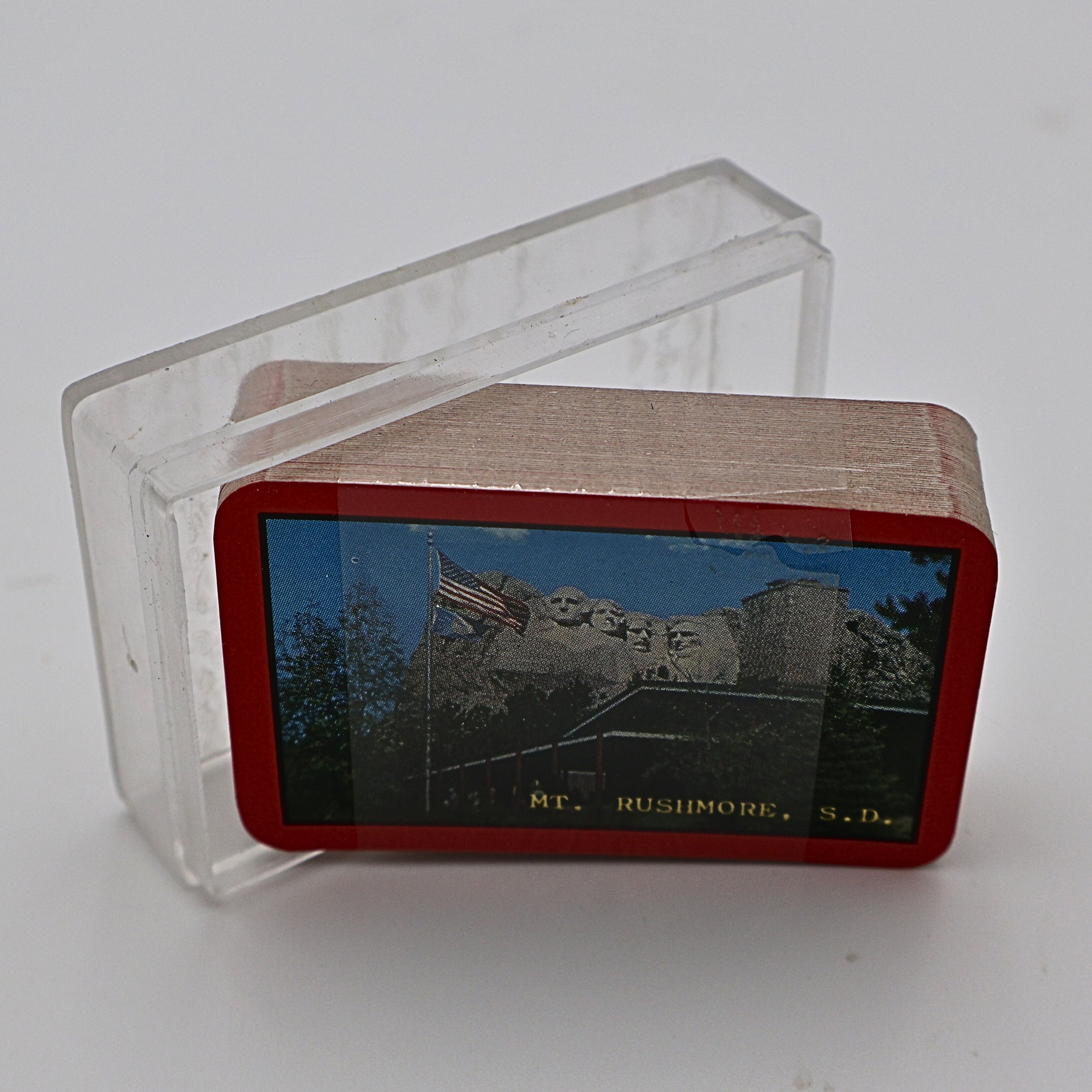 Vintage Mount Rushmore souvenir playing cards standing upright, showing the colorful Mount Rushmore National Memorial design printed on the card backs with a red border. The unopened travel souvenir deck remains sealed in its original plastic wrap and is photographed against a clean white background.