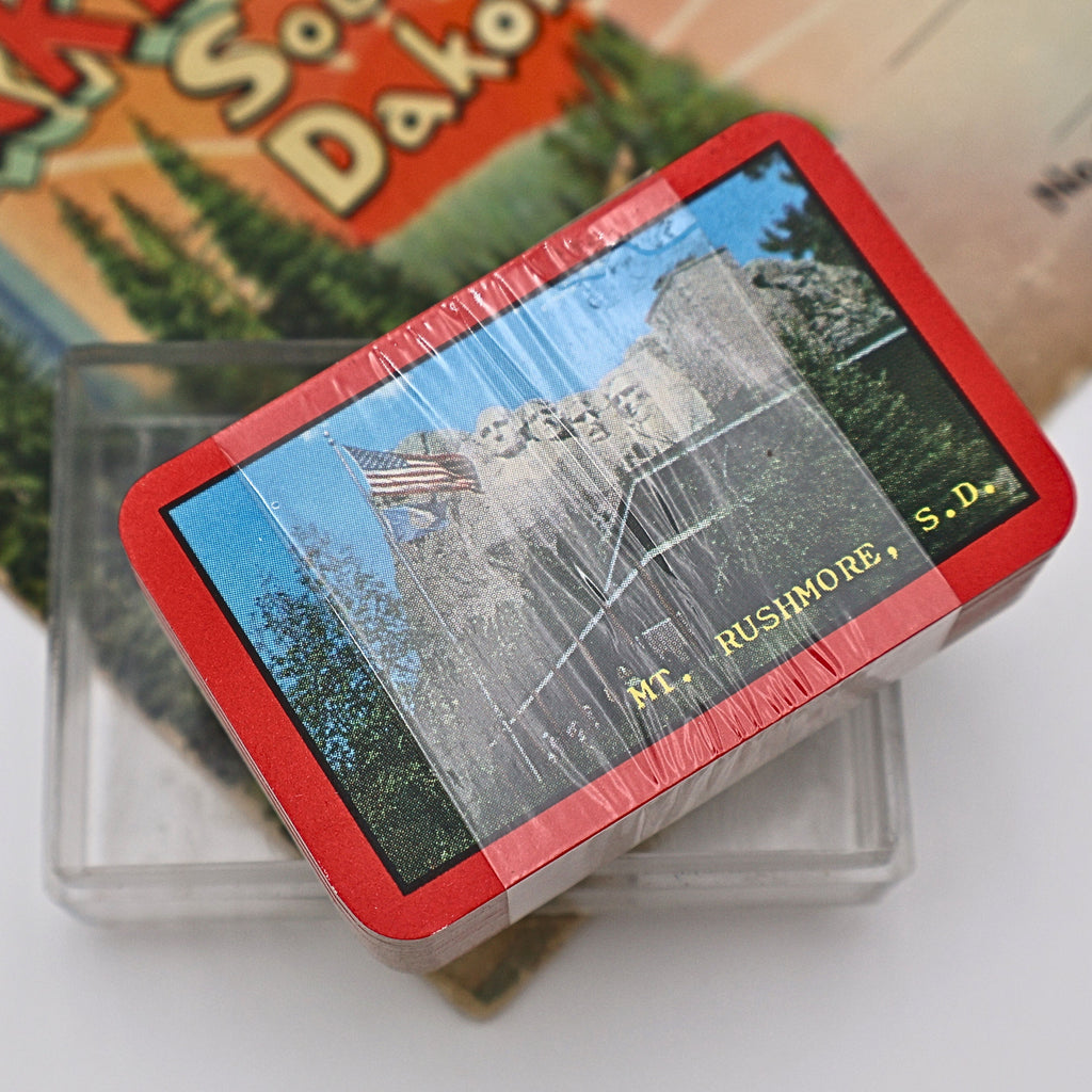 Close-up overhead view of the sealed vintage Mount Rushmore souvenir playing cards displaying the bright red border and scenic Mount Rushmore image. The original plastic wrap remains intact, highlighting this nostalgic travel collectible from South Dakota.