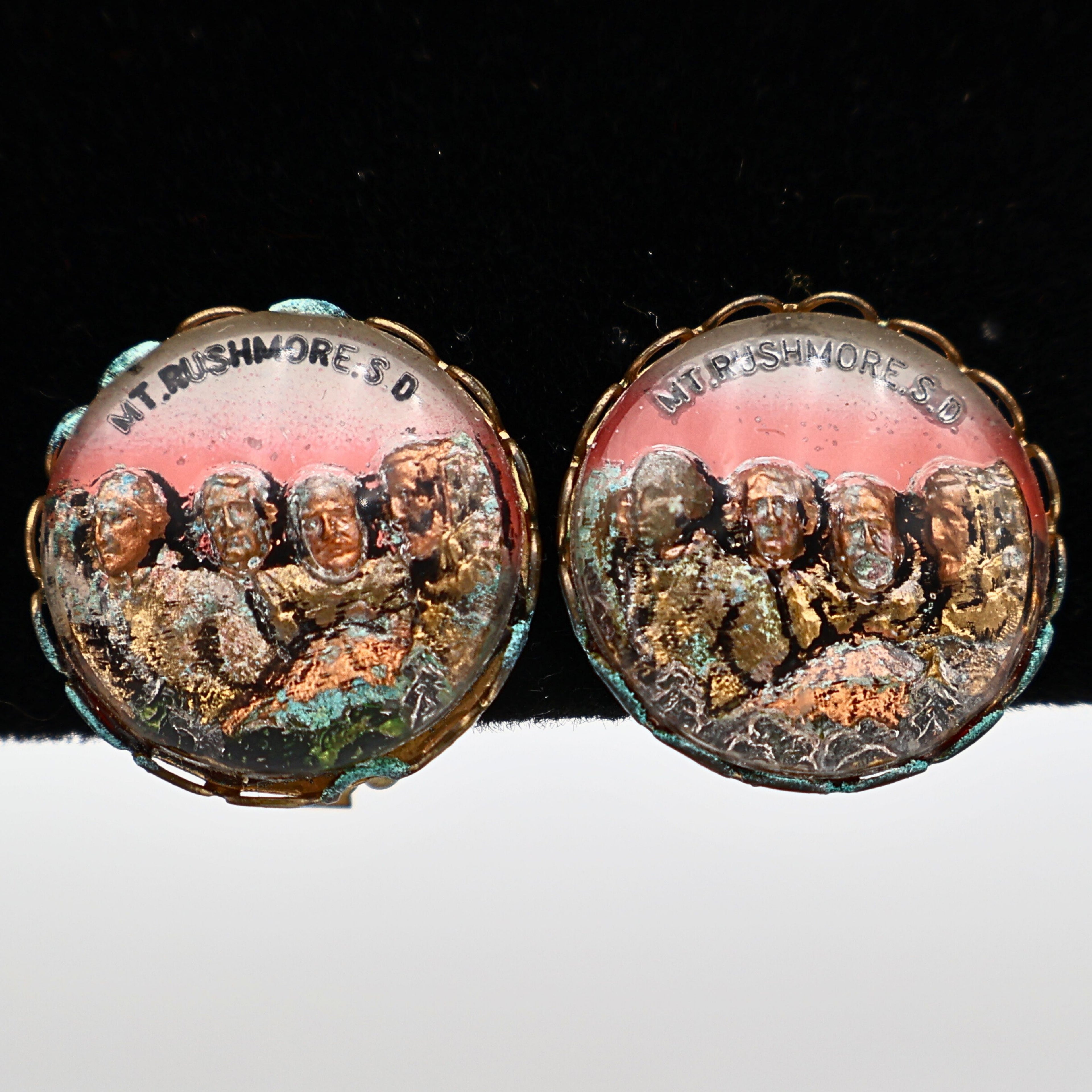 Pair of vintage Mount Rushmore clip-on earrings featuring raised sculpted presidential portraits beneath clear domed covers. The gold-tone scalloped settings frame colorful copper, bronze, green, and rose scenic details, highlighting classic mid-century South Dakota souvenir jewelry displayed from the front.
