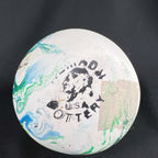 Underside of a Nemadji pottery vase showing a worn black Nemadji Pottery USA stamp with a profile illustration. Green and blue marbling crosses the pale circular base, with a rectangular tan patch on one side and several small marks against black fabric.