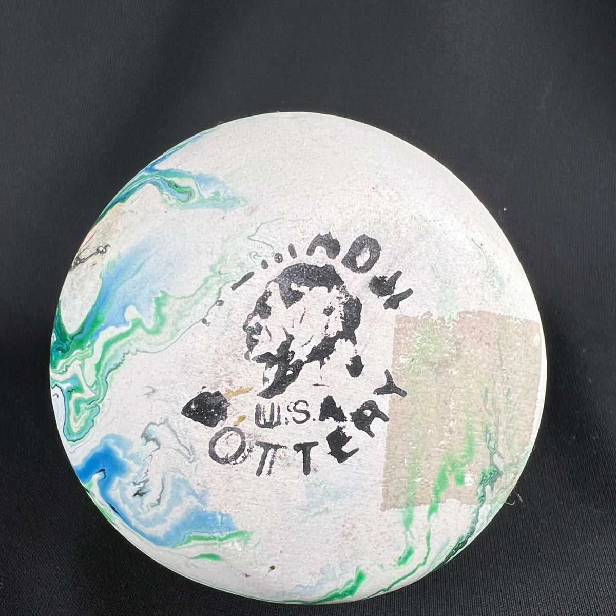 Underside of a Nemadji pottery vase showing a worn black Nemadji Pottery USA stamp with a profile illustration. Green and blue marbling crosses the pale circular base, with a rectangular tan patch on one side and several small marks against black fabric.