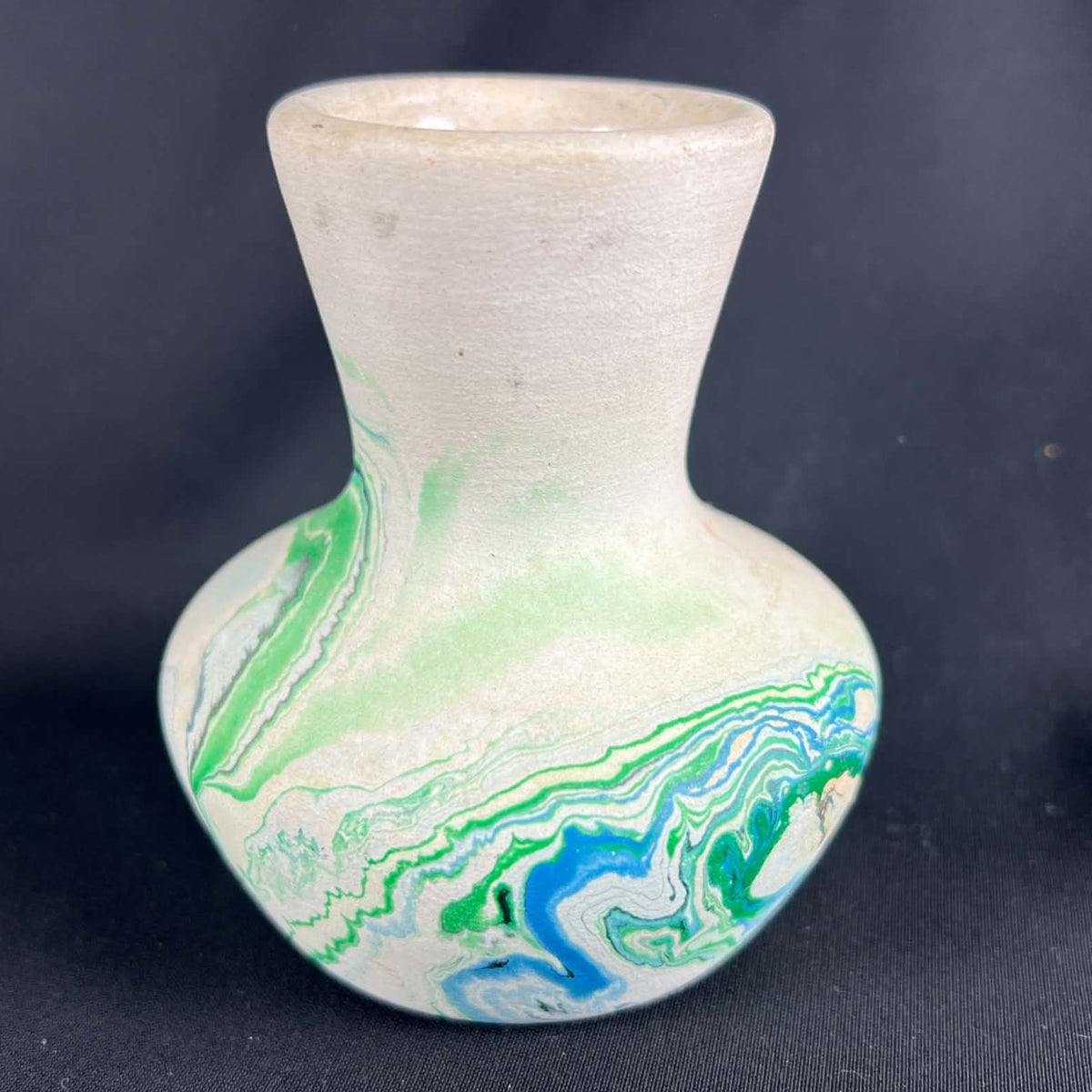 Nemadji pottery vase viewed from the side against black fabric, with a pale flared neck above a rounded body. Closely layered blue and green marbled ribbons curl across the lower half, while lighter green washes extend up the shoulder toward the neck.