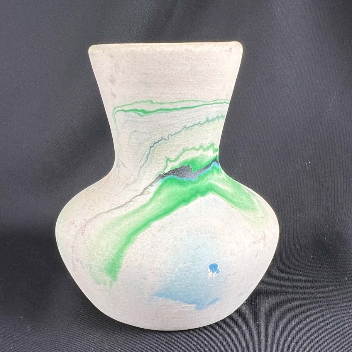 Side view of a Nemadji pottery vase with a rounded cream body, narrow neck and flared mouth on black fabric. A broad green band sweeps across the shoulder, outlined by fine gray and blue lines, with a pale blue patch and small blue spot lower on the body.