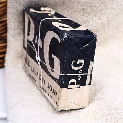 Three-quarter view of an original P&G White Laundry Soap bar in its vintage paper wrapper, displaying authentic age-related wear and classic Procter & Gamble branding. Perfect for vintage laundry room decor, advertising collections, primitive farmhouse displays, photography props, or nostalgic Americana decorating.