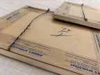 Angled rear view of the two frames, with one resting partly over the other. Twisted hanging wires, metal attachment loops, frame depth, brown backing paper, blue Jenson’s Upholstery tape, handwritten marks, and scattered age spots are visible.