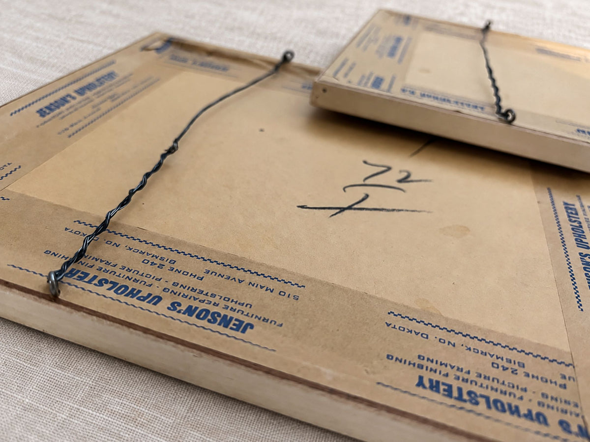 Angled rear view of the two frames, with one resting partly over the other. Twisted hanging wires, metal attachment loops, frame depth, brown backing paper, blue Jenson’s Upholstery tape, handwritten marks, and scattered age spots are visible.
