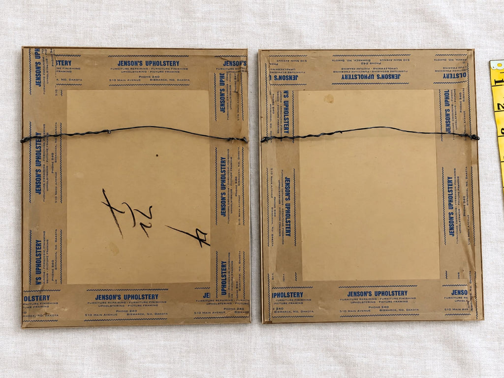 Rear view of both framed prints with brown paper backing, Jenson’s Upholstery printed tape, and attached wire hangers. One backing has large handwritten marks, and both show age-related spots. Part of a yellow measuring tape appears along the right edge.