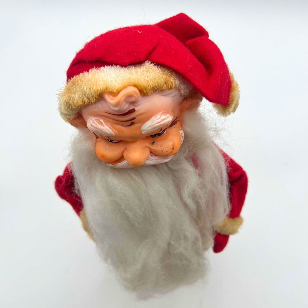 View from above of the vintage rubber face Santa doll, emphasizing his folded red cap, yellowed fuzzy hatband, modeled forehead curl, and white eyebrows. His sideways eyes, marked cheeks, curled mustache, and long white beard appear below the cap against a pale backdrop.