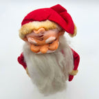 View from above of the vintage rubber face Santa doll, emphasizing his folded red cap, yellowed fuzzy hatband, modeled forehead curl, and white eyebrows. His sideways eyes, marked cheeks, curled mustache, and long white beard appear below the cap against a pale backdrop.
