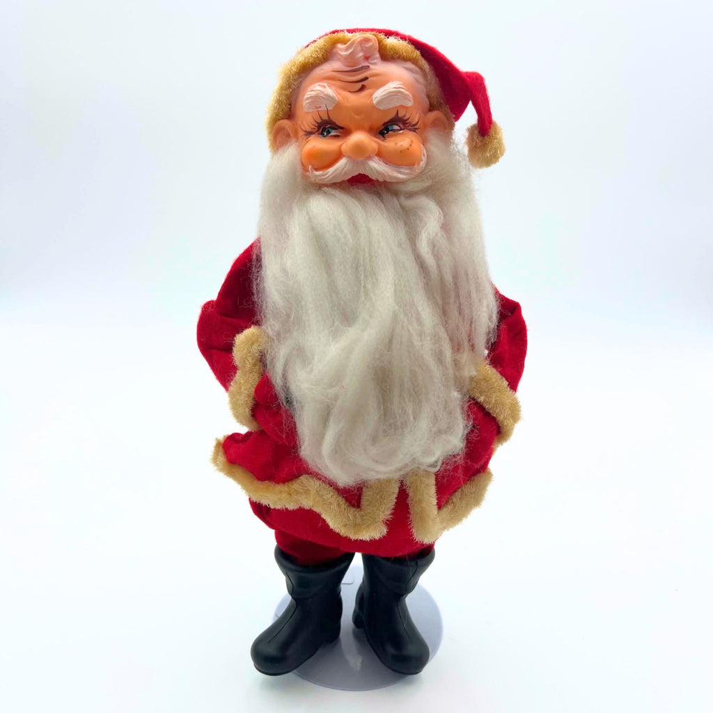 Front view of a vintage Sakai Santa Claus doll standing on a gray circular display base. His molded face has sideways-looking eyes and white eyebrows above a long white beard, with a red fabric suit, yellowed fuzzy trim, and black boots against a pale background.