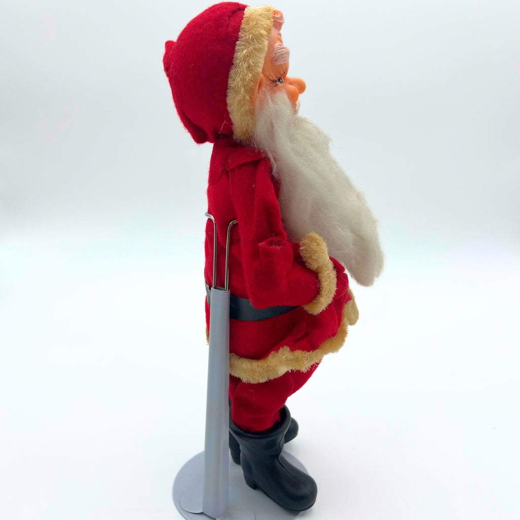 Right-facing side view of the vintage Sakai Santa Claus figure, with a long white beard extending over his red fabric coat. Yellowed fuzzy trim borders the cap, cuff, and hem; the black belt, molded boots, and gray metal display stand are visible behind him.