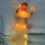 Front view of an illuminated vintage Christmas angel with gold-colored hair, a red bow, and outstretched silver mesh wings. Both raised candle lights glow amber, while colored bulbs shine through her flared mesh skirt beneath a cream bodice and silver belt.
