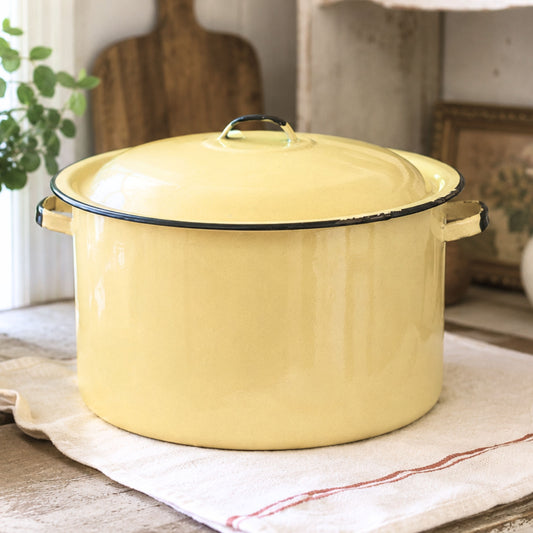 Vintage yellow enamelware stock pot with matching lid styled in a rustic farmhouse kitchen beside a sunny window. This large mid-century enamelware cooking pot features a buttery yellow finish with black trim and adds timeless charm to farmhouse, cottagecore, country, and vintage-inspired home decor.