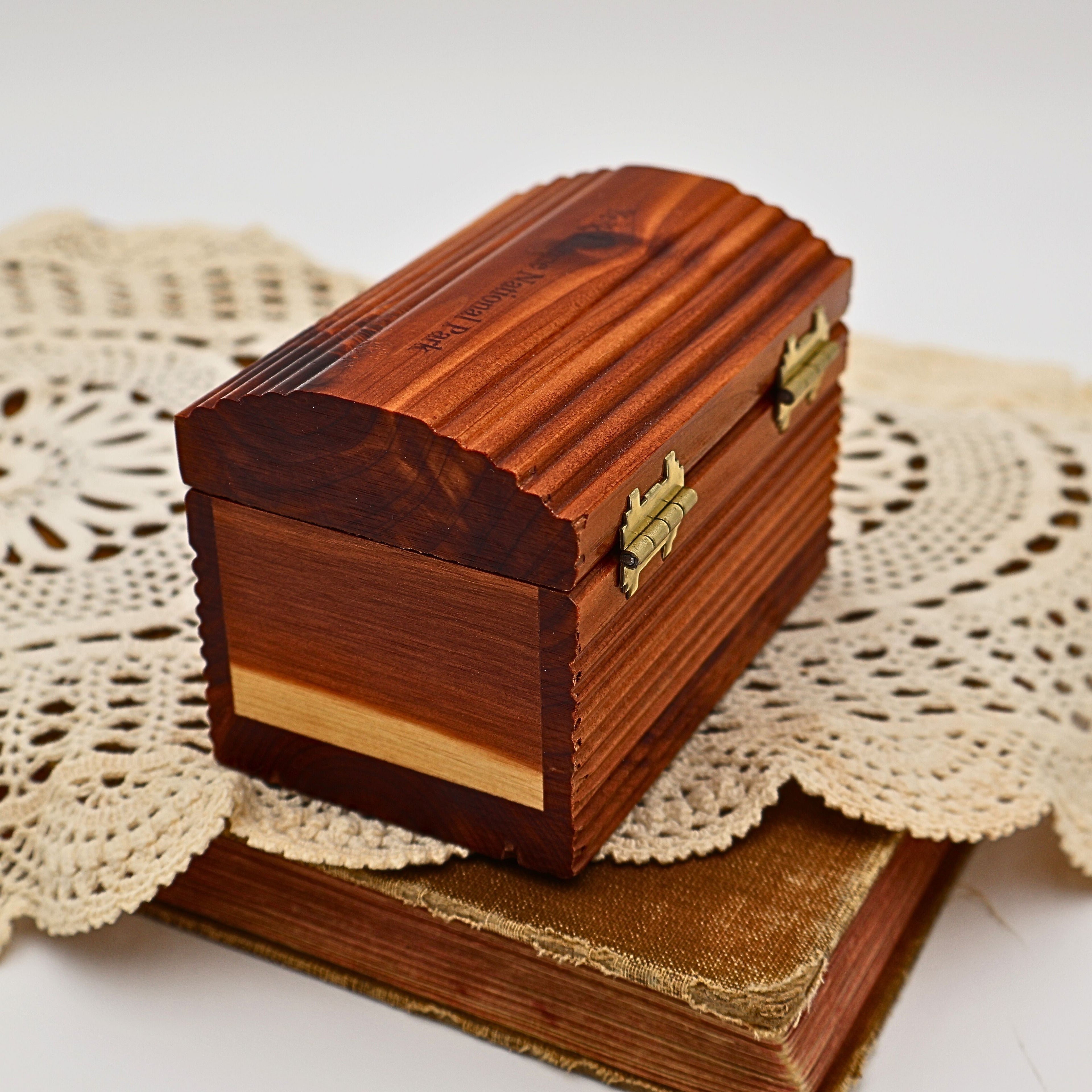 Side profile of the vintage Yellowstone National Park cedar keepsake box resting on antique books and a crocheted doily. Decorative brass-tone hinges, scalloped lid, and rich cedar grain showcase the timeless design of this handcrafted wooden souvenir box.