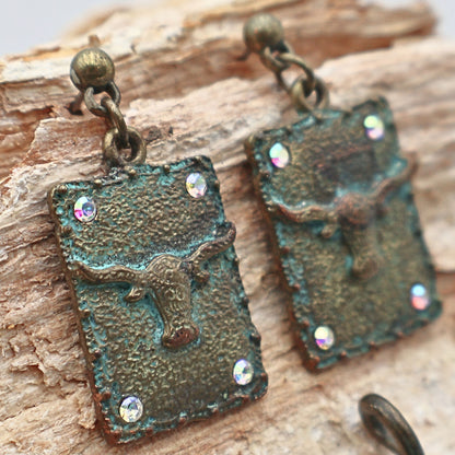 Close-up of Western longhorn dangle earrings with turquoise patina metal, ornate concho engraving, and rhinestone accents, lightweight rectangular cowgirl earrings with rustic texture and vintage-inspired finish, perfect for Western outfits, country concerts, rodeo fashion, and Southwestern boho jewelry styling