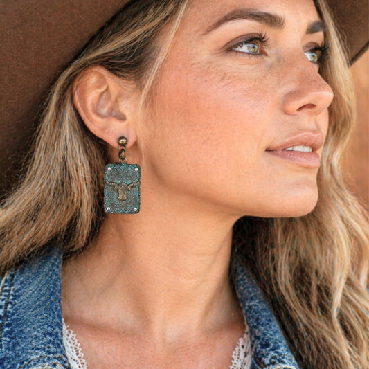 Western longhorn dangle earrings worn by model featuring turquoise patina finish with ornate concho detailing and rhinestone accents, cowgirl inspired statement jewelry styled with denim jacket and wide brim hat, rustic Western fashion accessory perfect for rodeo outfits, country concerts, boho cowgirl style, and Southwestern jewelry lovers